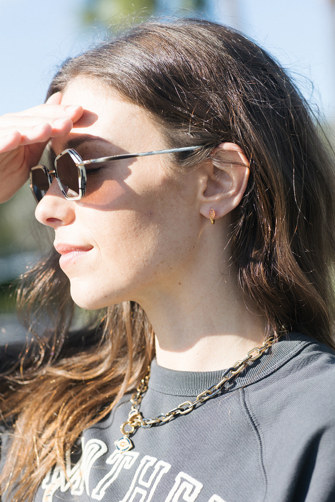 A close-up side profile of a woman wearing dark sunglasses with thin silver metal frames, a gold chain necklace, and a dark gray sweatshirt.