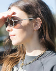 A close-up side profile of a woman wearing dark sunglasses with thin silver metal frames, a gold chain necklace, and a dark gray sweatshirt.