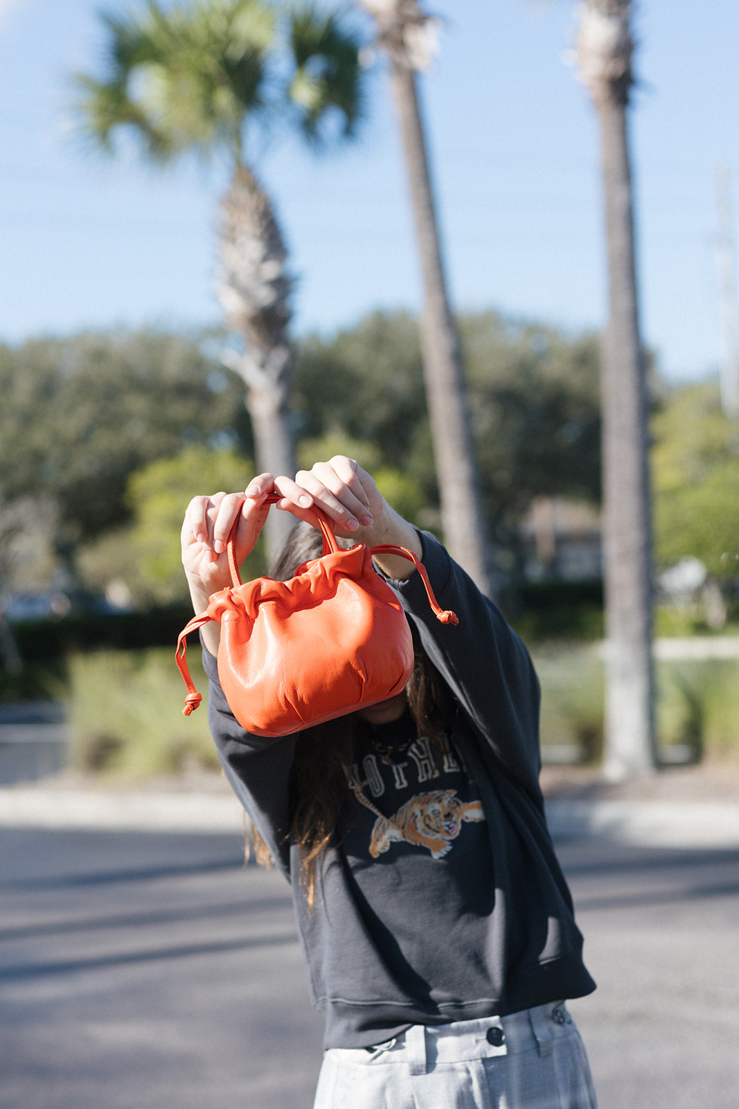 A person outdoors holding up a small orange drawstring pouch bag in front of their face, wearing a dark graphic sweatshirt and light denim jeans, with palm trees and a sunny street background.