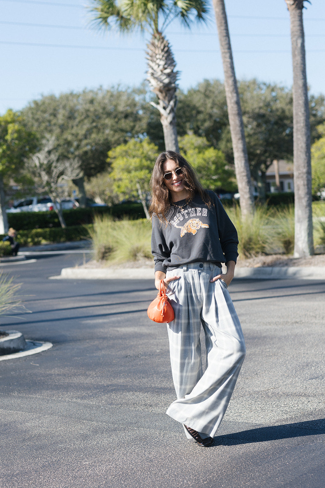 A woman walking outdoors on a sunny day wearing a dark gray graphic sweatshirt, light gray wide-leg plaid pants, and black shoes, holding an orange pumpkin. Palm trees and a street are visible in the background.