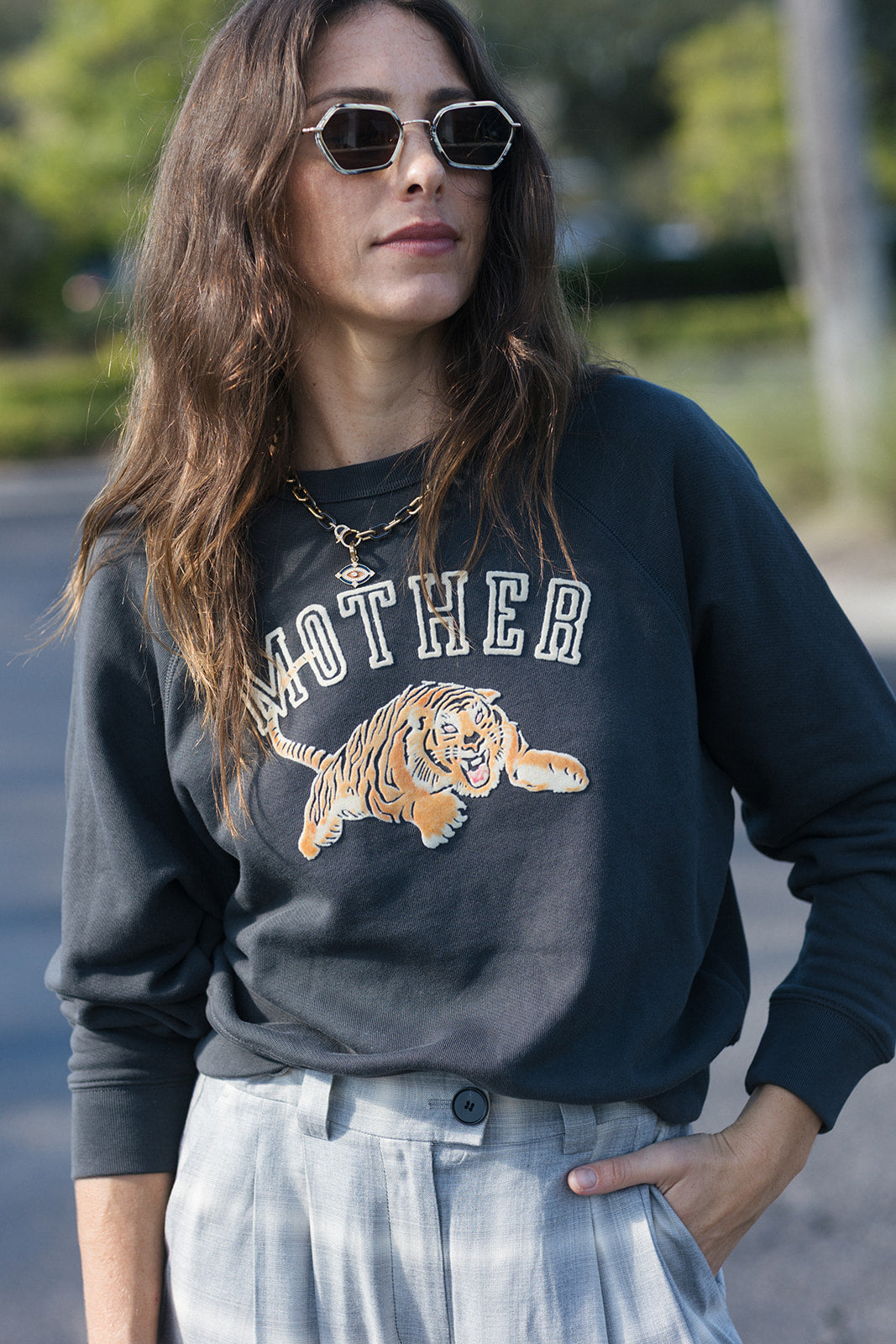 A woman wearing a dark charcoal gray sweatshirt with a graphic of a tiger and the word 'MOTHER' in white, paired with light blue jeans and sunglasses, photographed outdoors.