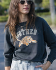 A woman wearing a dark charcoal gray sweatshirt with a graphic of a tiger and the word 'MOTHER' in white, paired with light blue jeans and sunglasses, photographed outdoors.