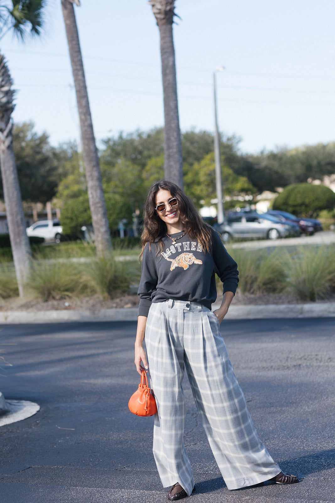 model wearing plaid pant with an orange bag and a sweatshirt in a parking lot