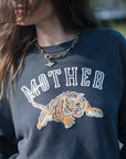 A close-up of a person wearing a navy blue sweatshirt with 'MOTHER' in white varsity letters and a large tiger graphic across the front. The person is also wearing a gold chain necklace.