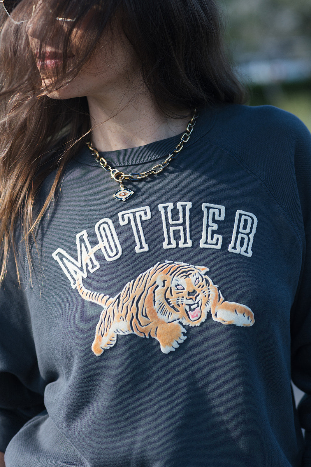 A close-up of a person wearing a navy blue sweatshirt with 'MOTHER' in white varsity letters and a large tiger graphic across the front. The person is also wearing a gold chain necklace.