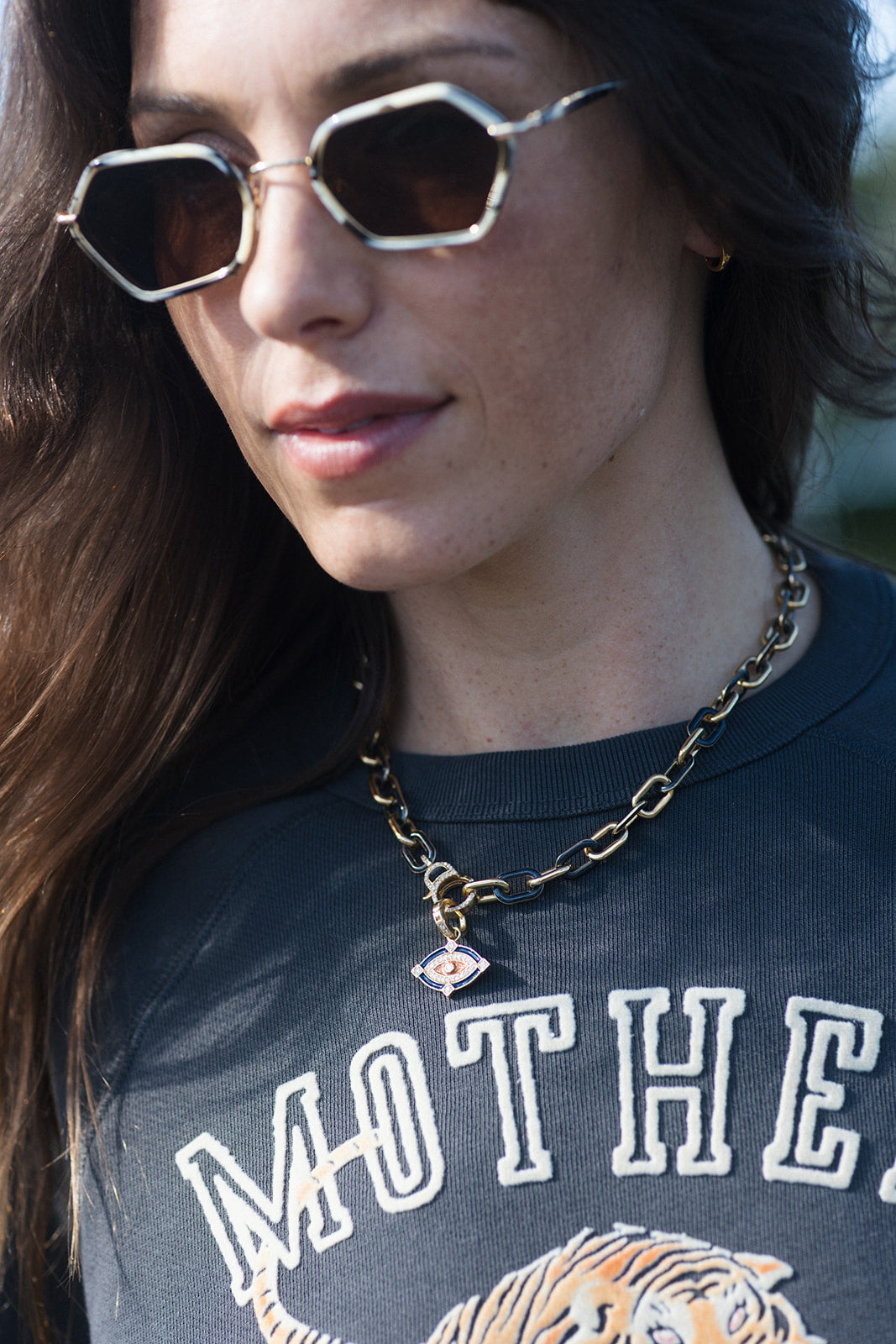 A close-up lifestyle shot of a woman wearing a gold chain necklace with a small evil eye pendant, paired with geometric sunglasses and a dark graphic t-shirt.
