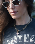 A close-up lifestyle shot of a woman wearing a gold chain necklace with a small evil eye pendant, paired with geometric sunglasses and a dark graphic t-shirt.
