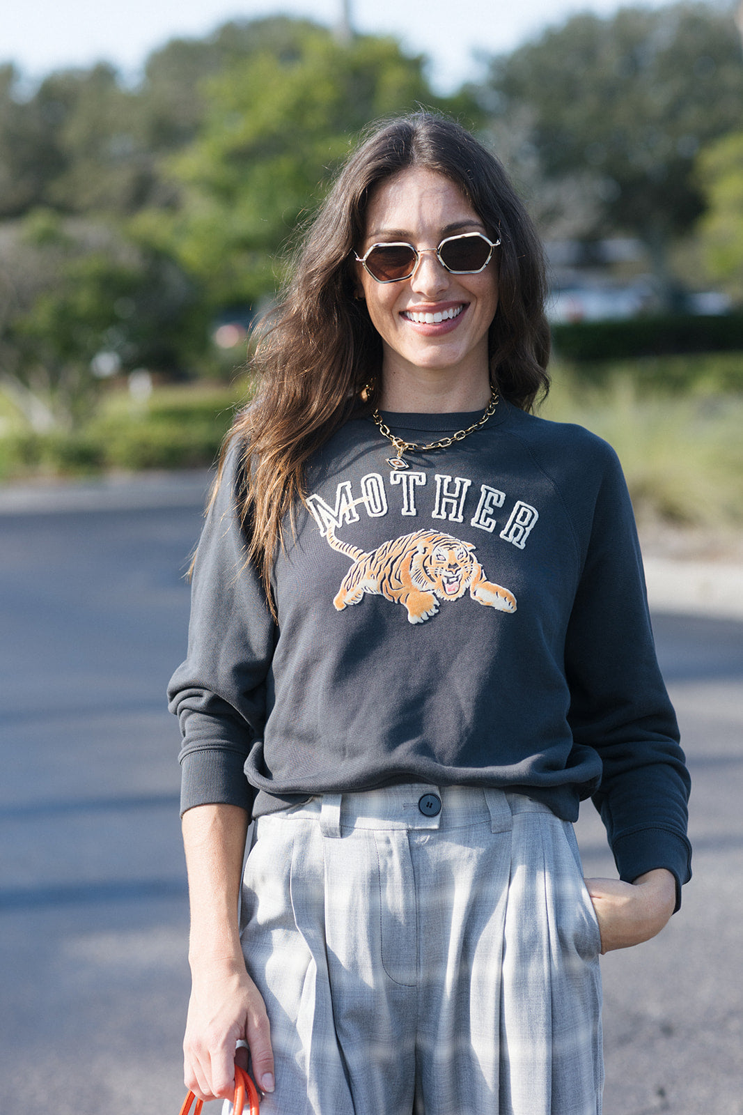 A woman wearing a dark charcoal gray crewneck sweatshirt with a graphic of a tiger and the word 'MOTHER' in white and orange, paired with light gray tie-dye pants, standing outdoors on a paved path.