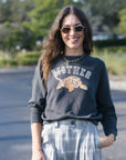 A woman wearing a dark charcoal gray crewneck sweatshirt with a graphic of a tiger and the word 'MOTHER' in white and orange, paired with light gray tie-dye pants, standing outdoors on a paved path.