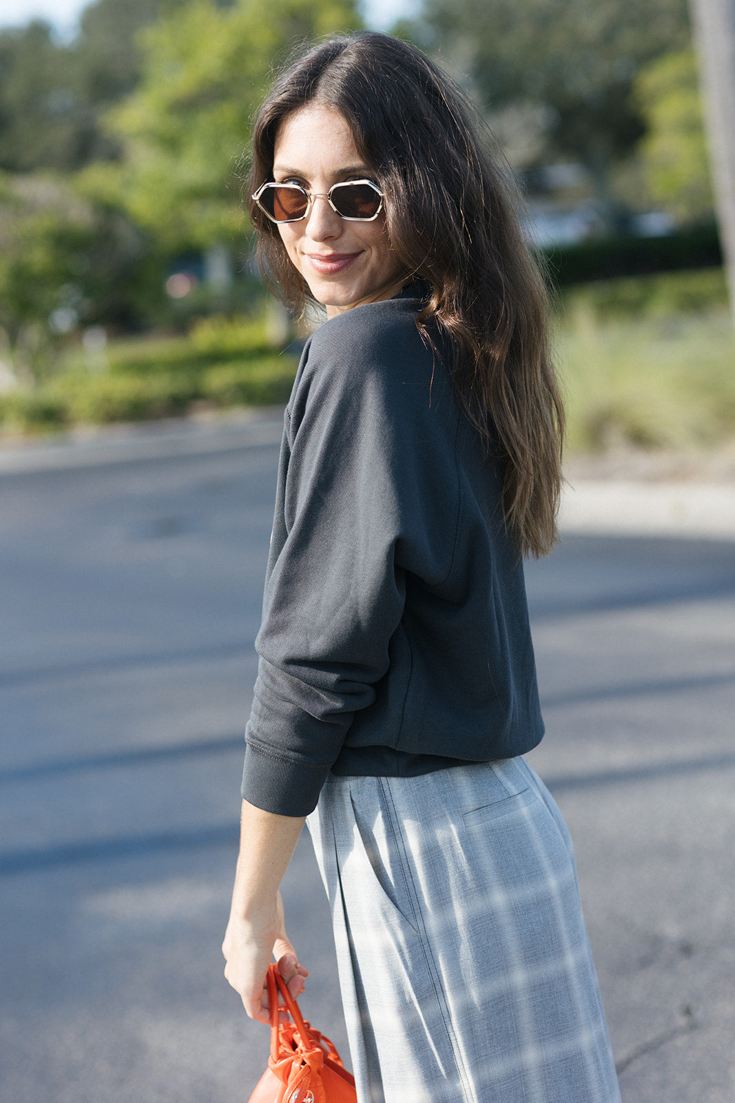 A woman wearing a dark charcoal gray long-sleeve crewneck sweatshirt and a light gray and white plaid skirt, standing outdoors on a paved road with trees in the background.