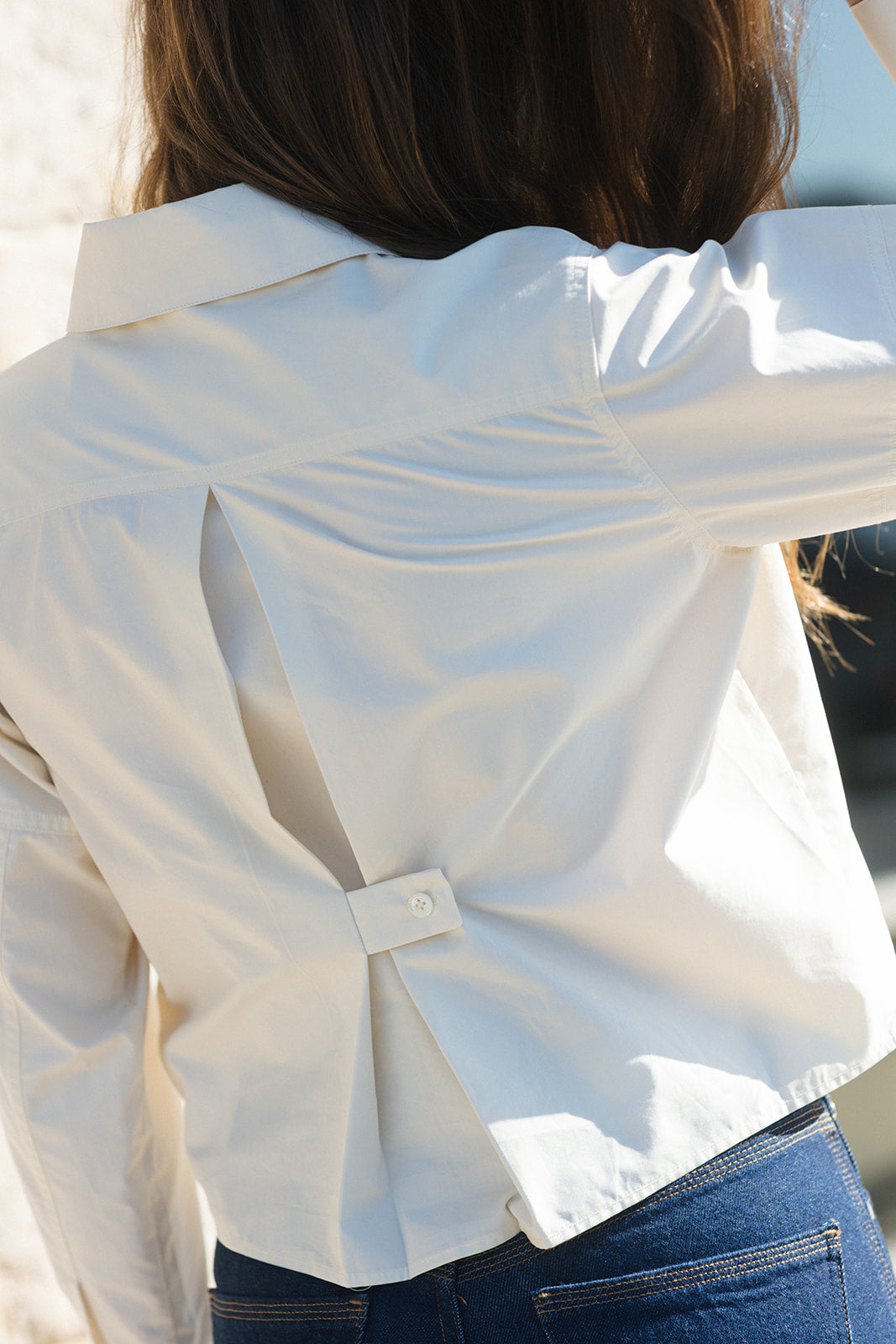 A close-up back view of a person wearing a white button-front shirt with a unique split back detail and blue jeans.
