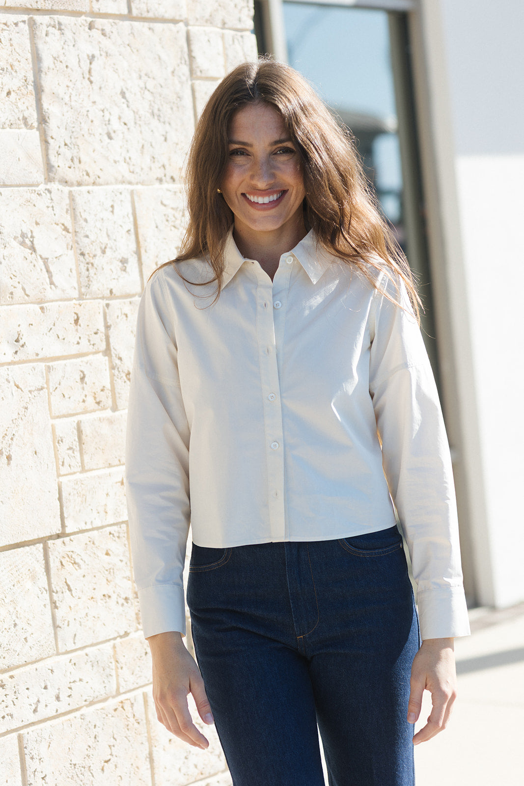 A woman with long brown hair smiling, wearing a white long-sleeve button-down shirt and dark blue jeans, standing outdoors against a light beige stone wall.