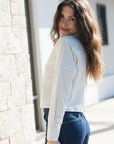 A woman with long brown hair is shown from the back wearing a white long-sleeve button-up shirt and dark blue jeans, standing outdoors next to a light-colored stone wall.