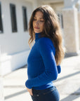 A woman with long brown hair wearing a vibrant royal blue ribbed turtleneck sweater and dark blue jeans, standing outdoors in bright sunlight against a white building.