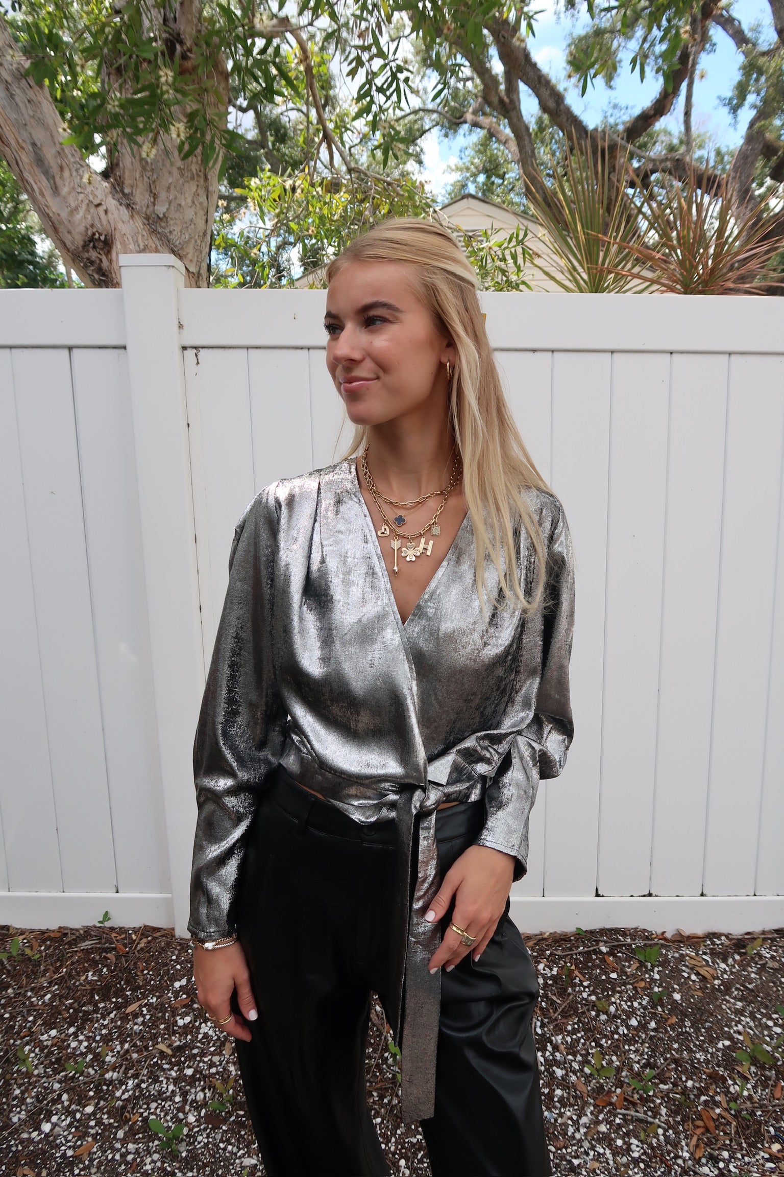 Woman wearing a silver metallic top and black pants standing outdoors with a white fence and trees in the background.