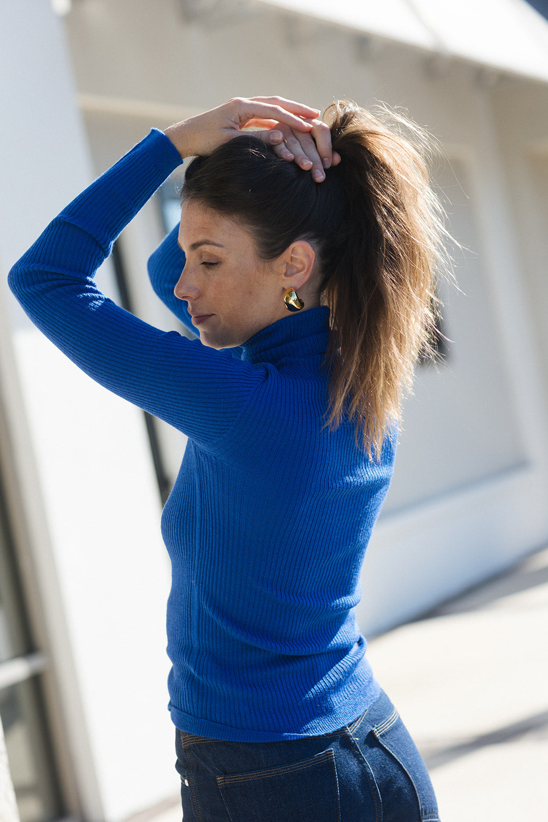 A woman wearing a bright blue ribbed turtleneck sweater and dark jeans, adjusting her hair in a sunny outdoor setting.