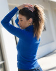 A woman wearing a bright blue ribbed turtleneck sweater and dark jeans, adjusting her hair in a sunny outdoor setting.