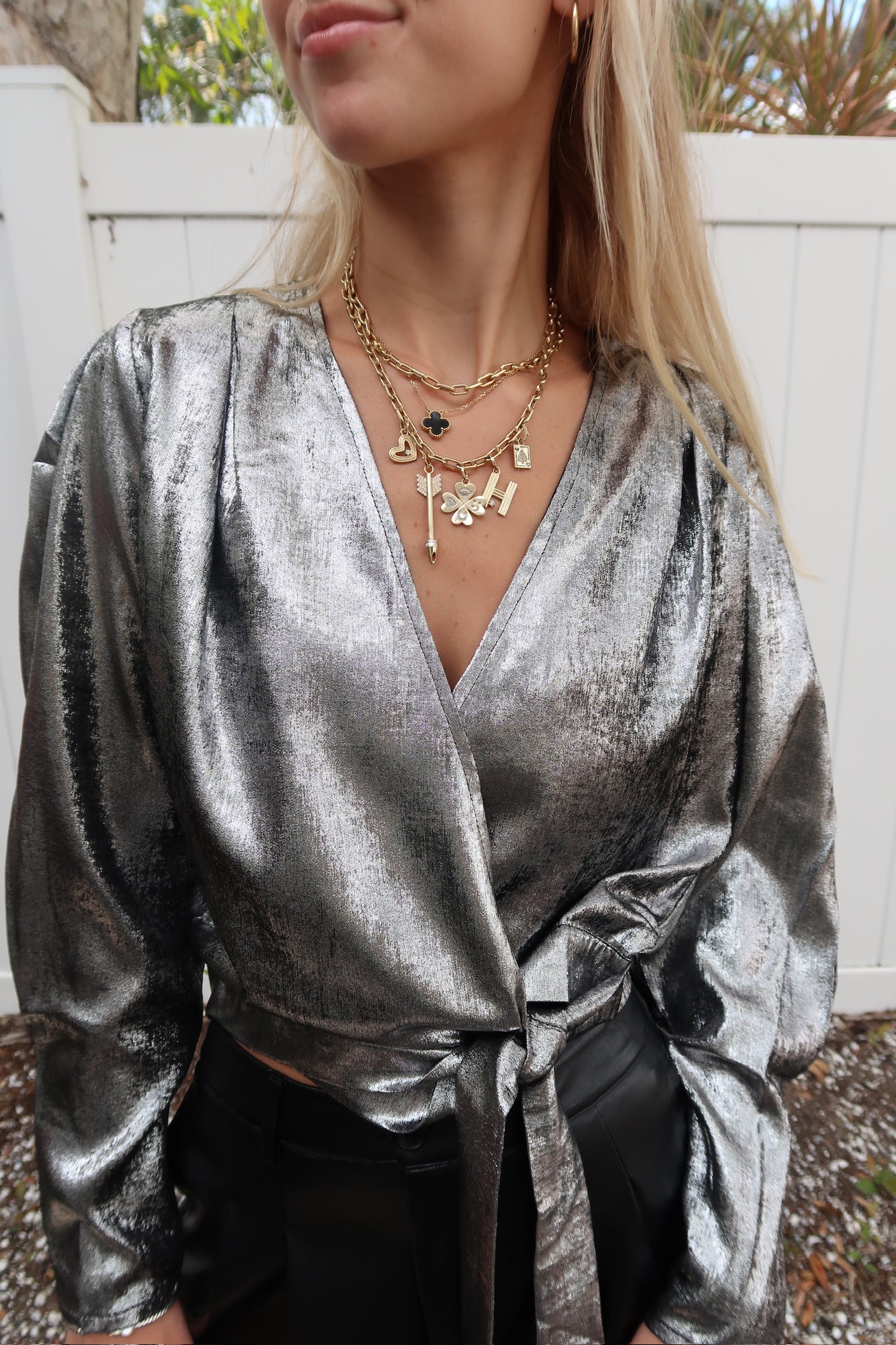 Person wearing a silver metallic top with layered necklaces against a white fence background, close up