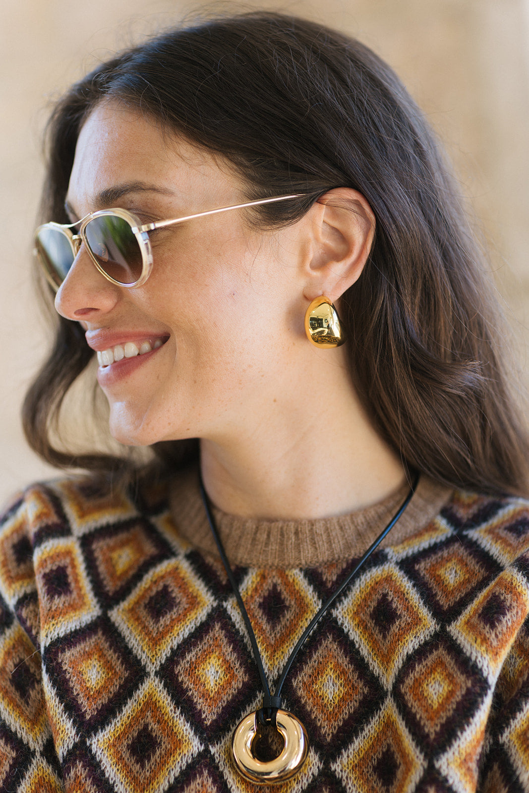 A woman in profile wearing a geometric patterned knit sweater, gold hoop earrings, and a gold pendant necklace, set against a warm beige background.