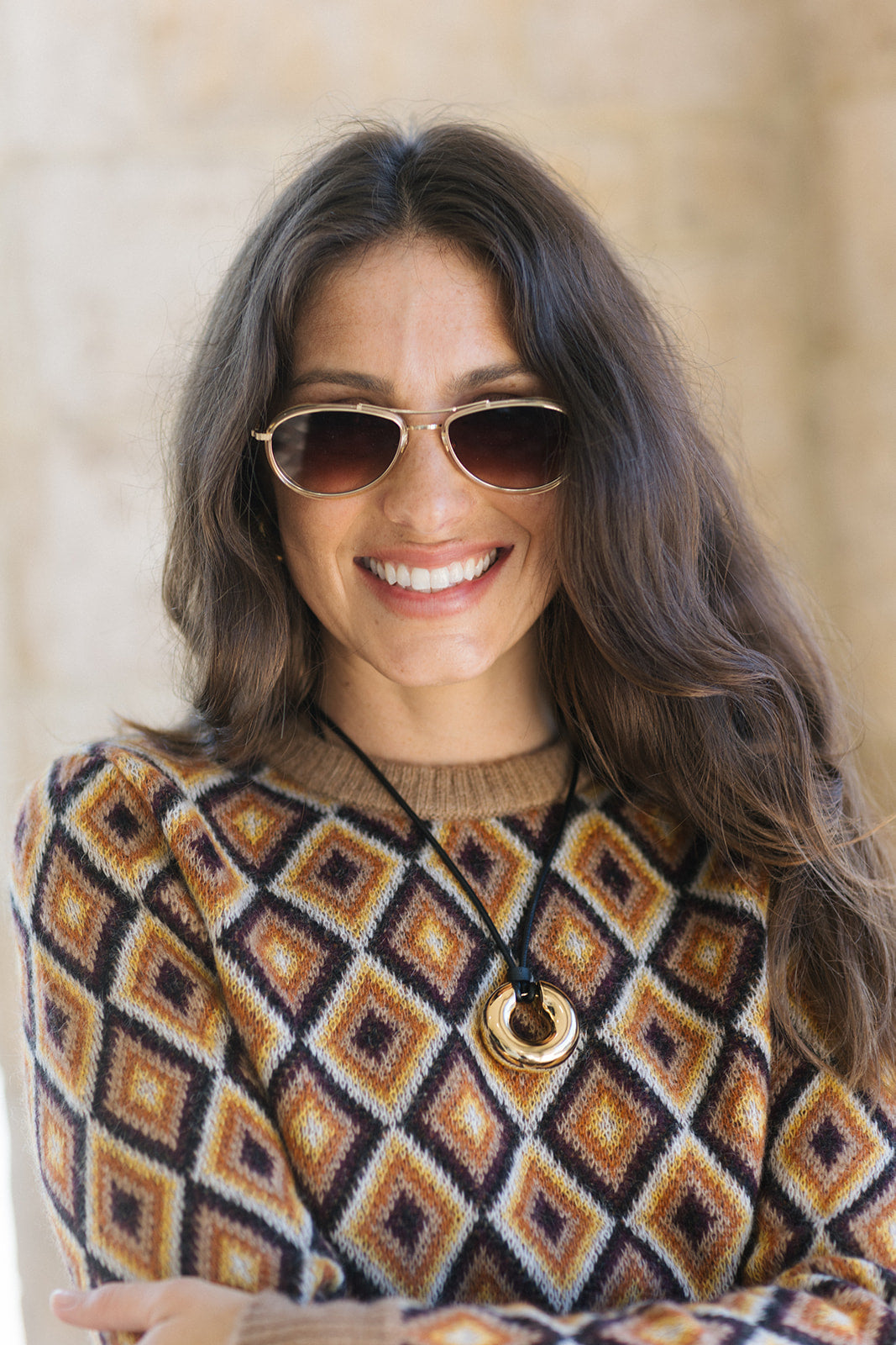 A woman with long brown hair wearing a brown and beige geometric patterned knit sweater, gold-framed sunglasses, and a gold pendant necklace, smiling against a soft beige background.
