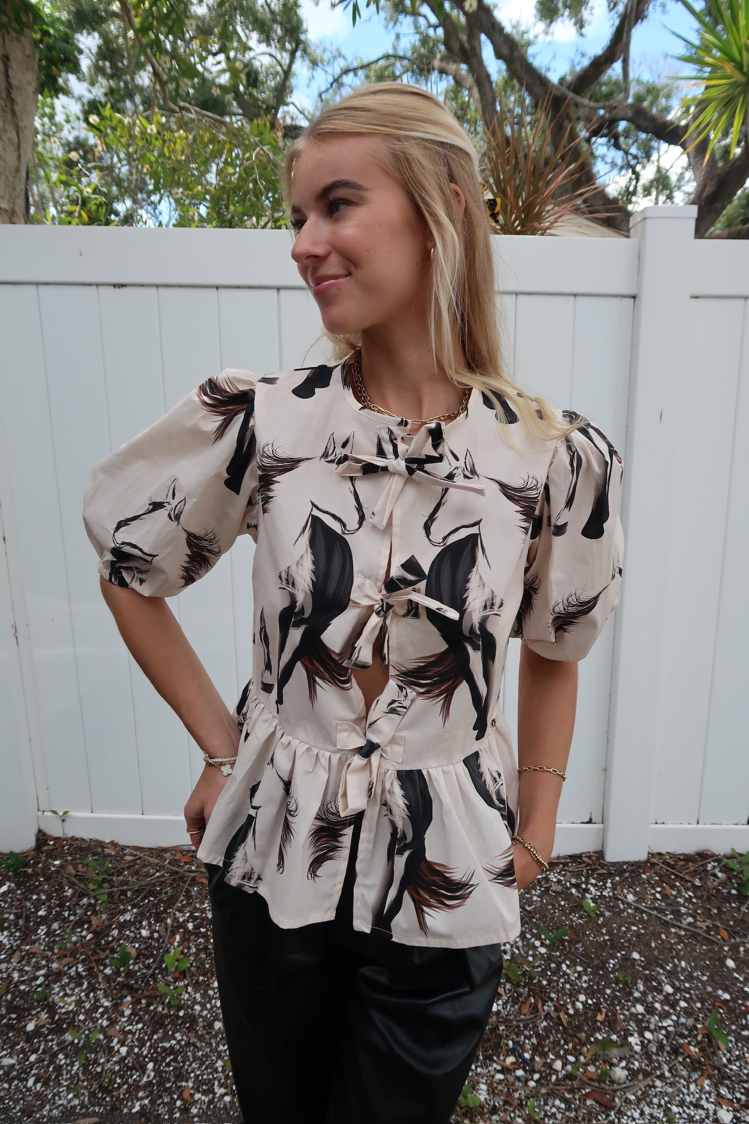 Woman wearing a patterned blouse standing outdoors with a white fence and trees in the background