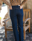 A person viewed from behind wearing dark blue straight-leg jeans, a brown and beige diamond-patterned knit sweater, standing on a stone patio next to a wooden bench.