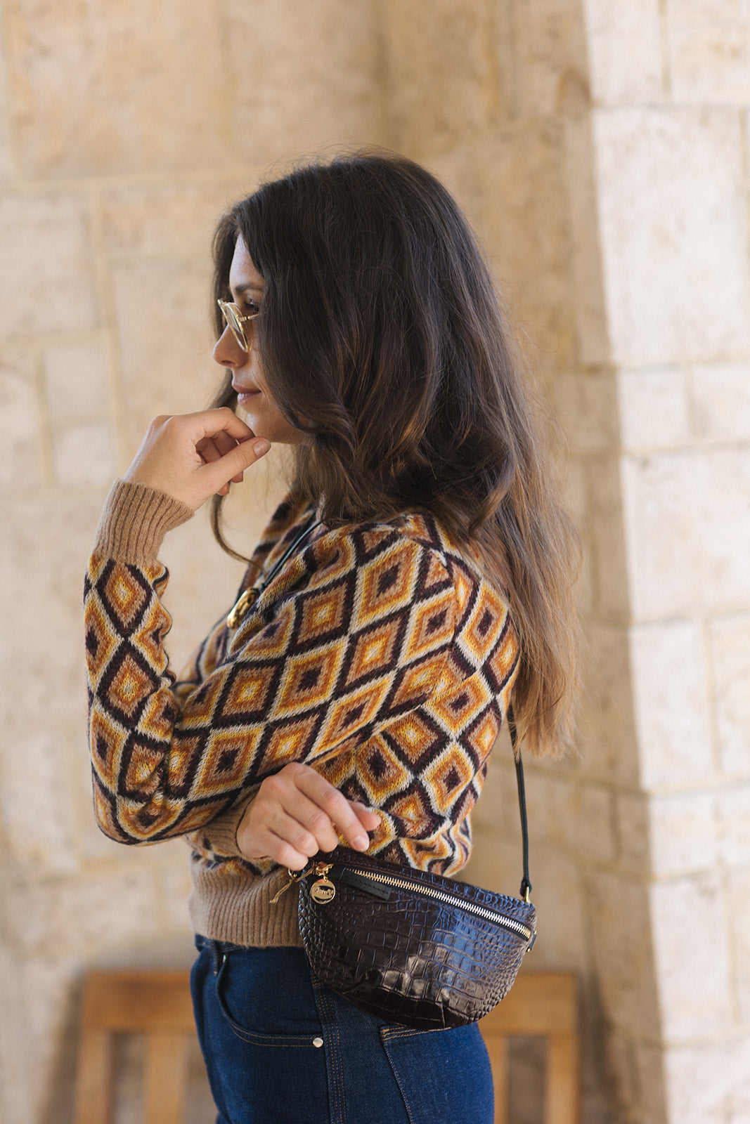 A woman with long dark hair and glasses is shown in profile, wearing a brown, beige, and black diamond-patterned knit sweater, blue jeans, and carrying a small black textured shoulder bag.