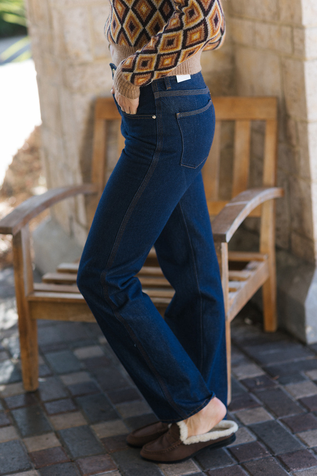 A person wearing dark blue straight-leg jeans, a brown and beige diamond-patterned knit sweater, and brown slippers, standing next to a wooden bench in an outdoor patio setting.