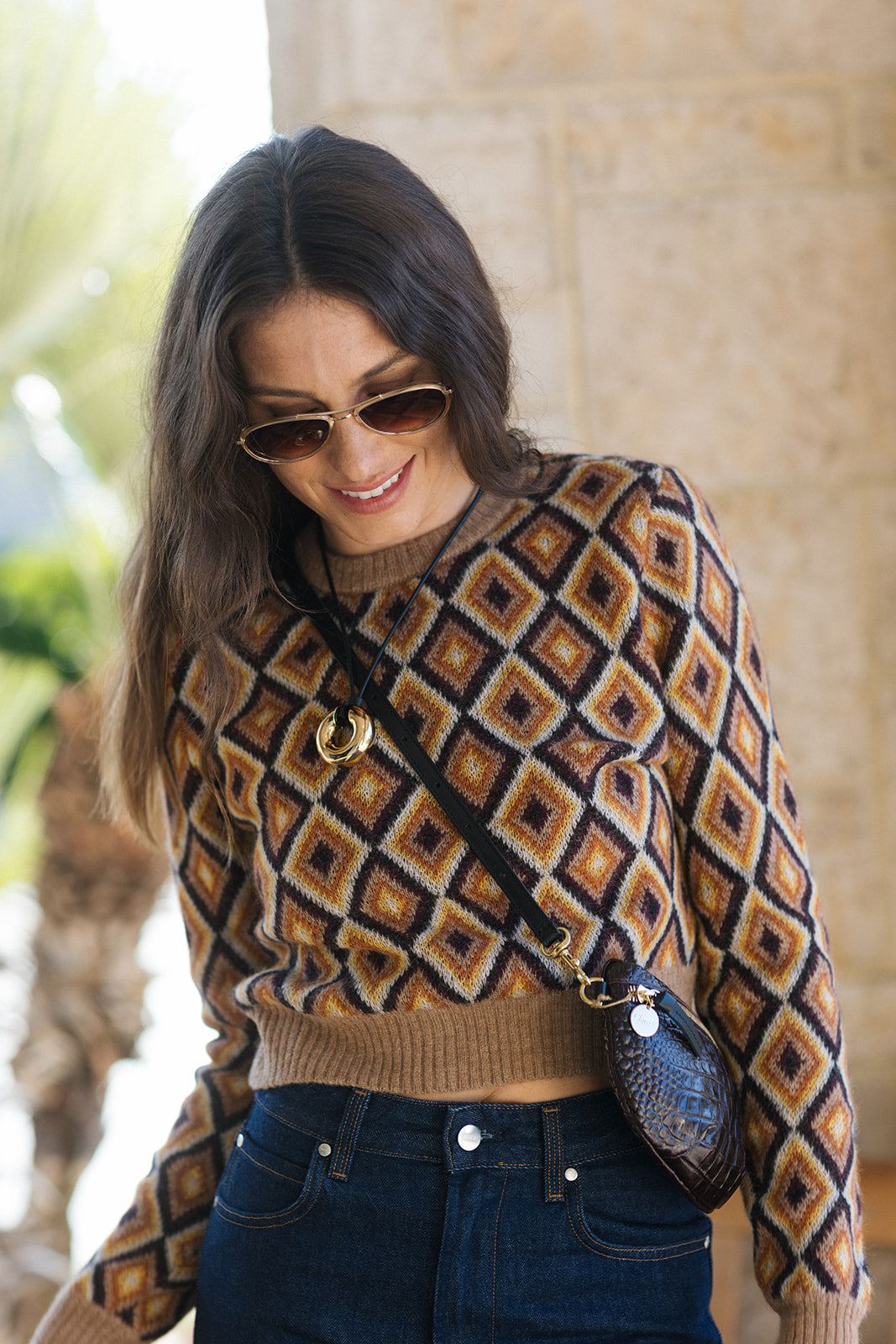 A woman wearing a brown, beige, and black geometric diamond-patterned knit sweater, dark blue jeans, and sunglasses, carrying a small snakeskin-patterned crossbody bag.