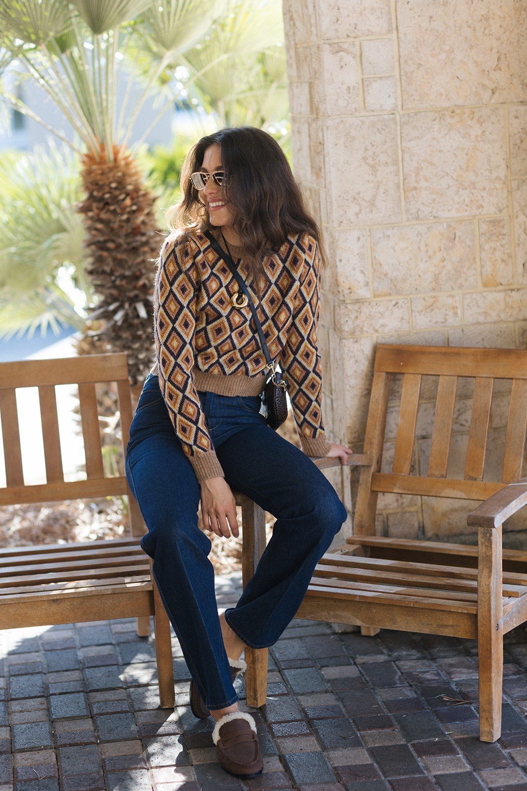 A woman wearing a brown and beige diamond-patterned knit crop top, dark blue jeans, and brown loafers sits on a wooden bench in an outdoor patio setting.