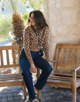 A woman wearing a brown and beige diamond-patterned knit crop top, dark blue jeans, and brown loafers sits on a wooden bench in an outdoor patio setting.