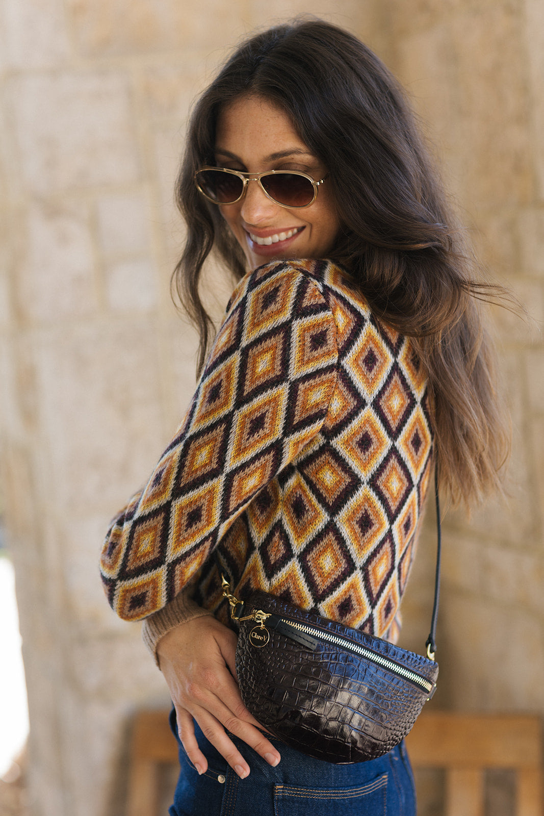 A woman wearing a brown, orange, and cream geometric diamond-patterned knit sweater, blue jeans, and gold-framed sunglasses, carrying a dark textured crocodile-embossed crossbody bag.