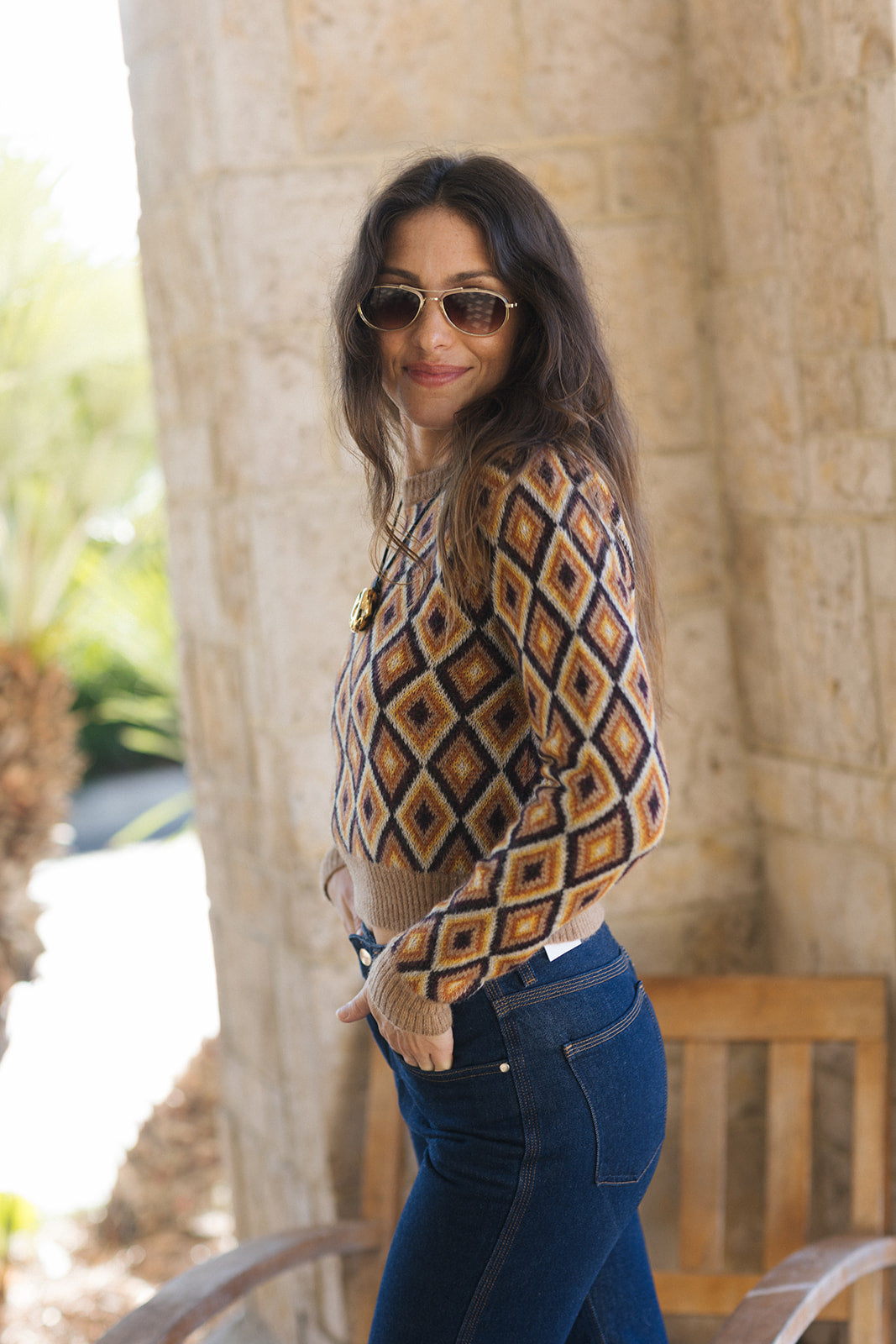 A woman wearing a brown, beige, and navy geometric diamond-pattern knit sweater and blue jeans, standing outdoors in front of a stone column.