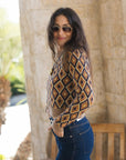 A woman wearing a brown, beige, and navy geometric diamond-pattern knit sweater and blue jeans, standing outdoors in front of a stone column.