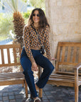 A woman wearing a brown and beige geometric patterned long-sleeve crop top, dark blue jeans, and brown slip-on shoes sits on a wooden bench in an outdoor patio setting.