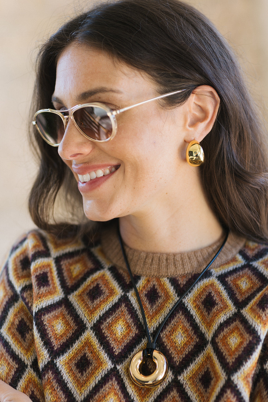 A woman wearing gold hoop earrings with a small central gold bead, styled with a geometric brown, beige, and black knit sweater and gold-framed sunglasses.