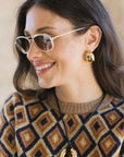 A woman wearing gold hoop earrings with a small central gold bead, styled with a geometric brown, beige, and black knit sweater and gold-framed sunglasses.