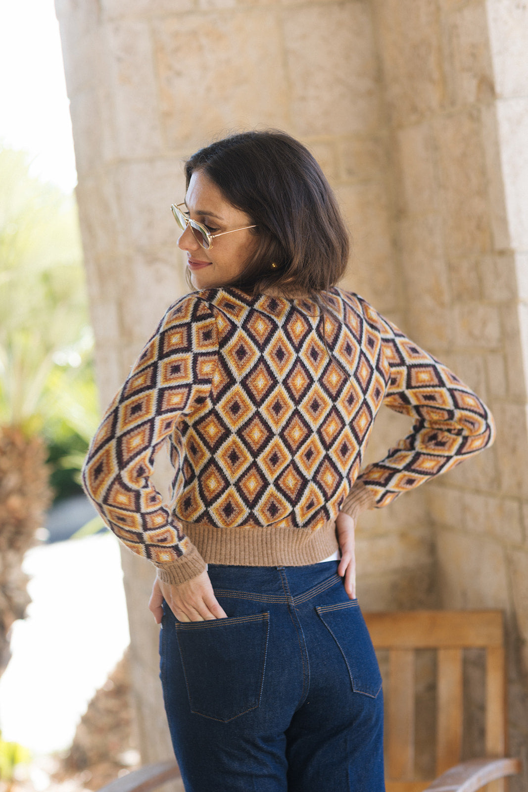 A woman wearing a cropped long-sleeve knit sweater with a geometric diamond pattern in beige, brown, and navy, paired with dark blue jeans, photographed outdoors against a stone pillar.