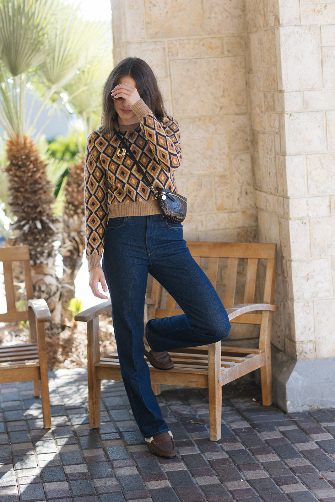 A woman posing outdoors in a brown and beige diamond-patterned knit sweater, dark blue jeans, and brown shoes, carrying a small black crossbody bag.