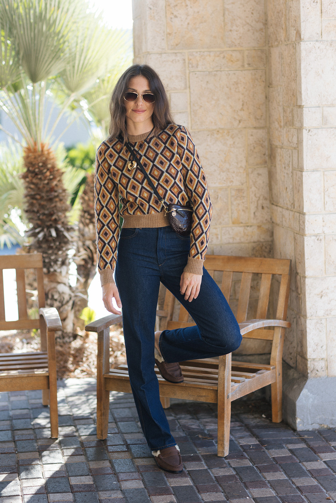 A woman posing outdoors in a patterned brown and beige long-sleeve crop top, dark blue jeans, and brown loafers, sitting on a wooden bench.