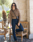 A woman posing outdoors in a patterned brown and beige long-sleeve crop top, dark blue jeans, and brown loafers, sitting on a wooden bench.