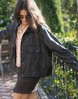 A woman wearing an oversized brown leather jacket and a dark brown mini skirt, photographed from behind in an outdoor garden patio setting.
