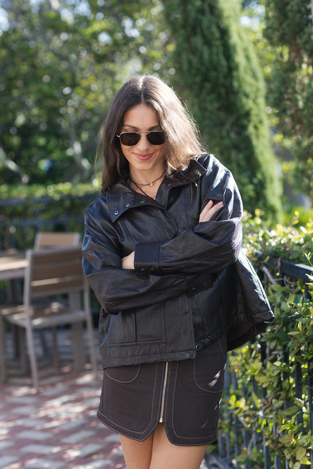 A woman wearing an oversized brown leather jacket and a dark brown mini skirt, photographed from behind in an outdoor garden patio setting.