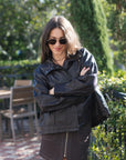 A woman wearing an oversized brown leather jacket and a dark brown mini skirt, photographed from behind in an outdoor garden patio setting.