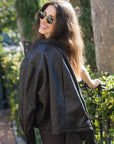A woman wearing a brown leather jacket and sunglasses, smiling and looking back over her shoulder in an outdoor garden setting.