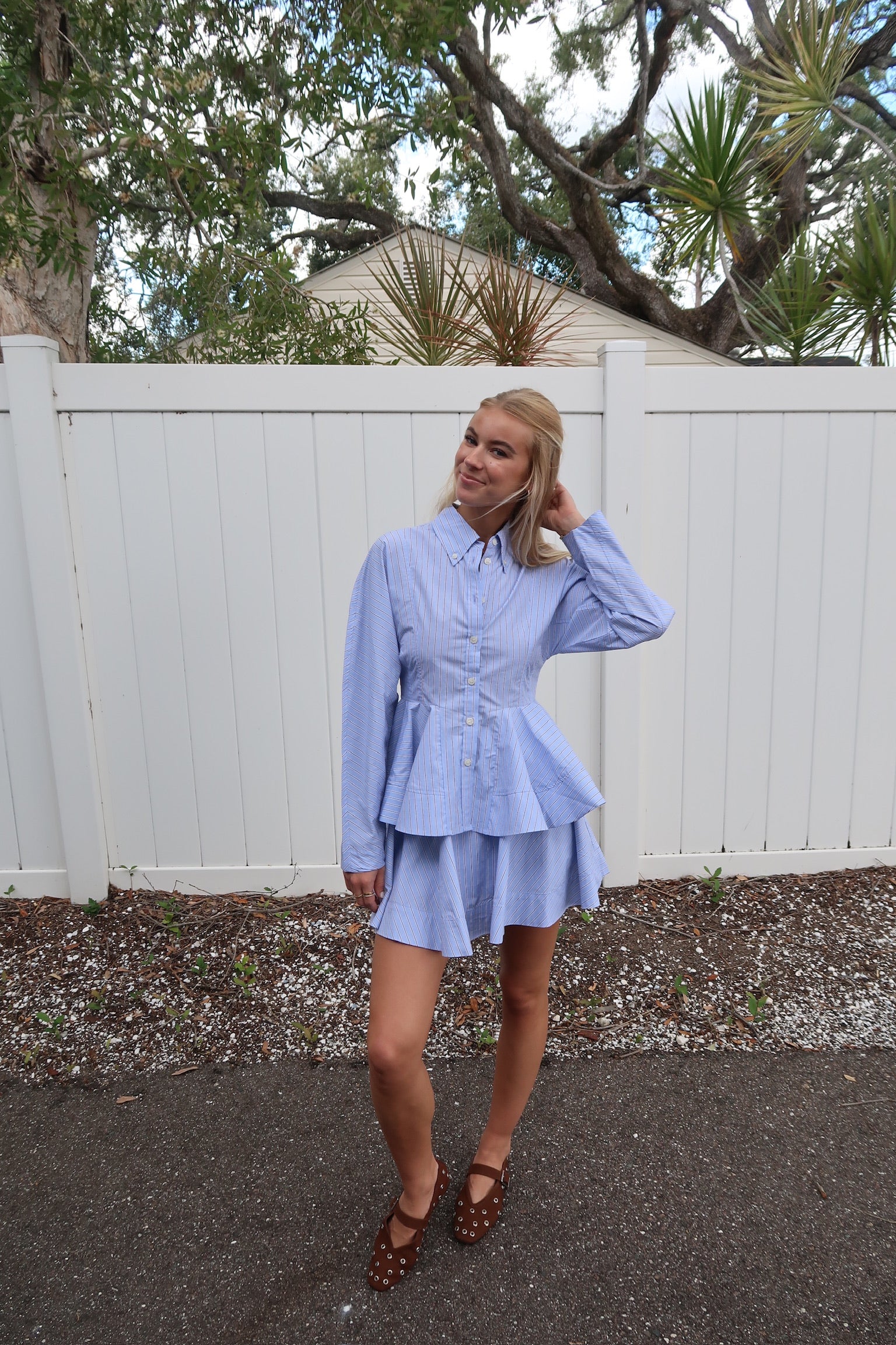 Woman wearing a light blue set standing in front of a white fence with trees in the background
