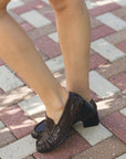 A person wearing dark brown textured slip-on loafers with low block heels, standing on a red and beige brick pavement outdoors.