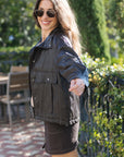 A woman wearing a brown utility jacket  and sunglasses, posing outdoors in a garden setting.