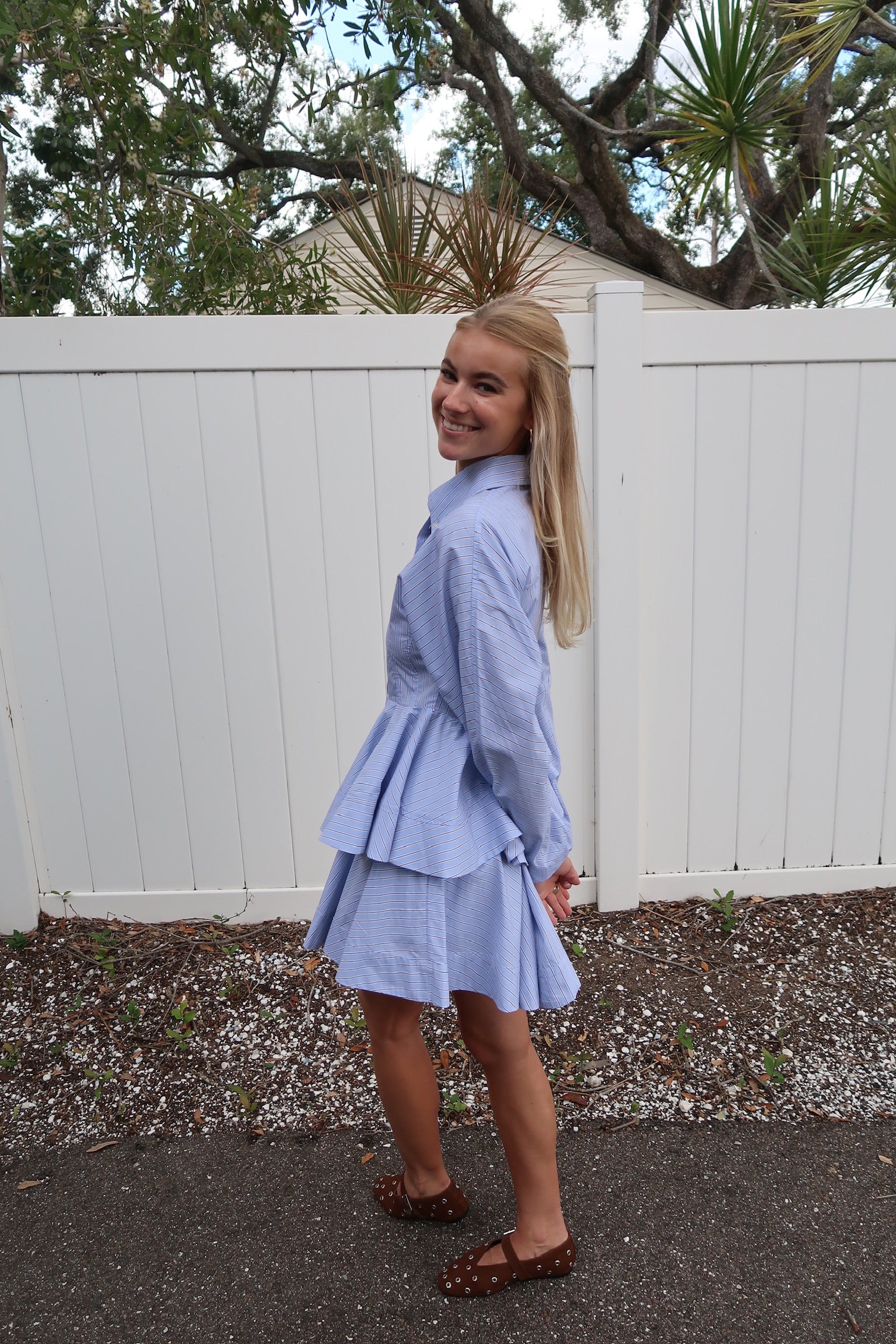 Young girl in a light blue set standing outdoors with a white fence and trees in the background, side view