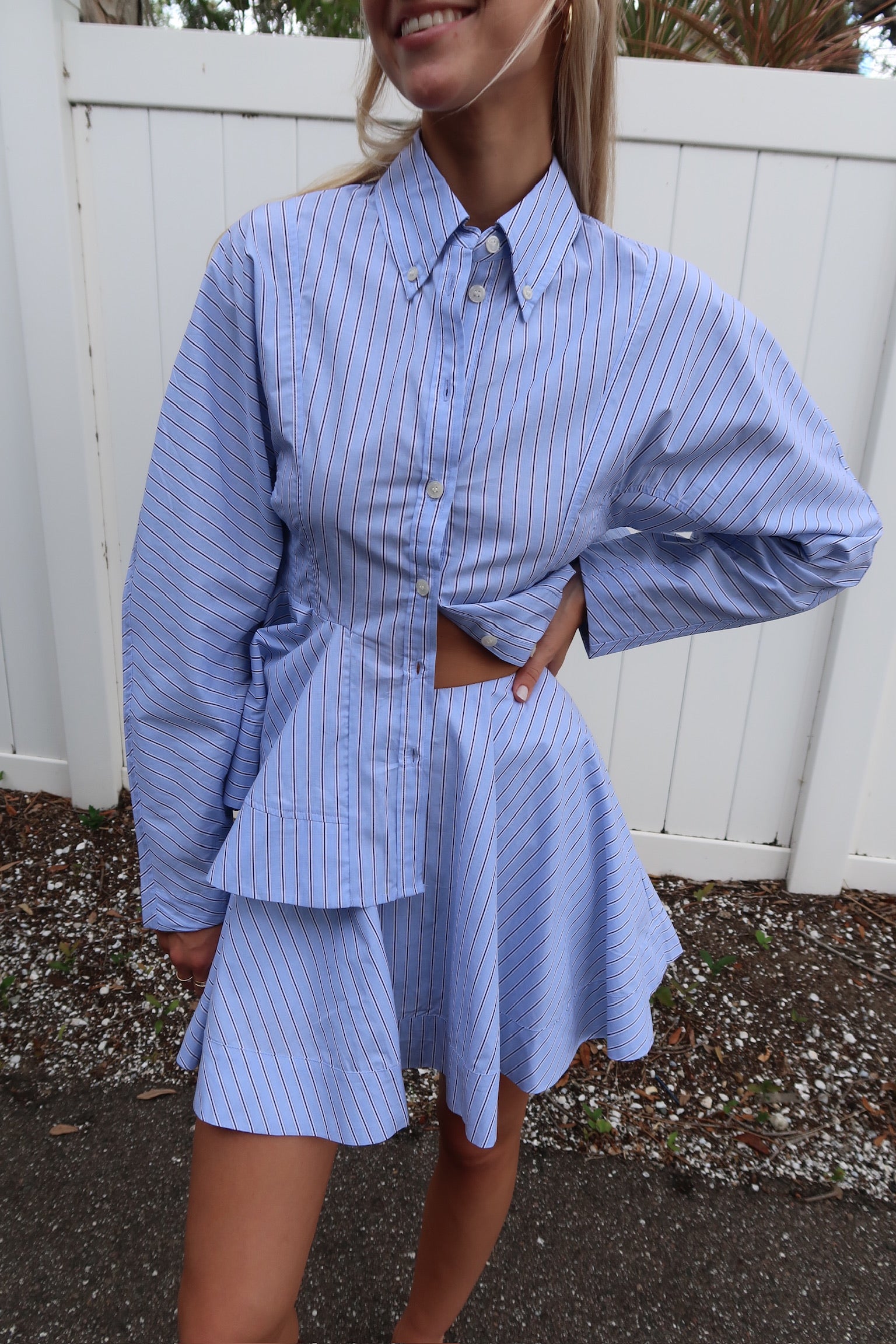 Person wearing a blue striped shirt and skirt set against a white fence.